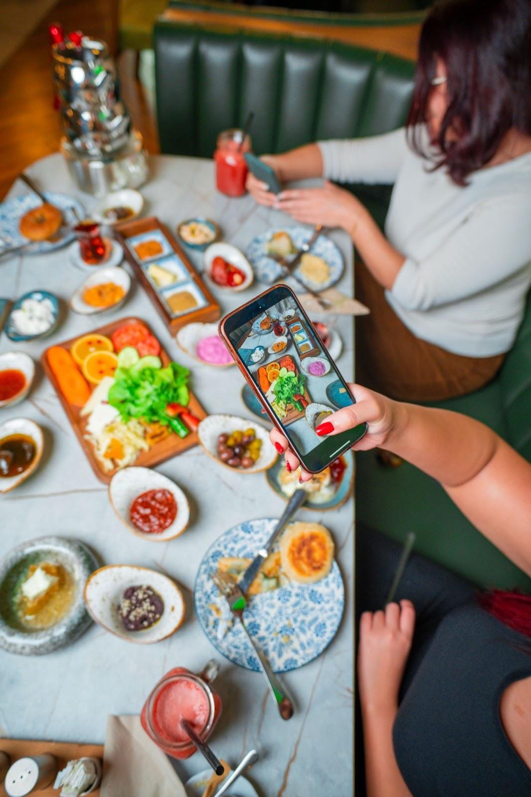Traditional Turkish breakfast spread with assorted cheeses, jams, olives, and bread, as people enjoy and photograph the colorful dishes