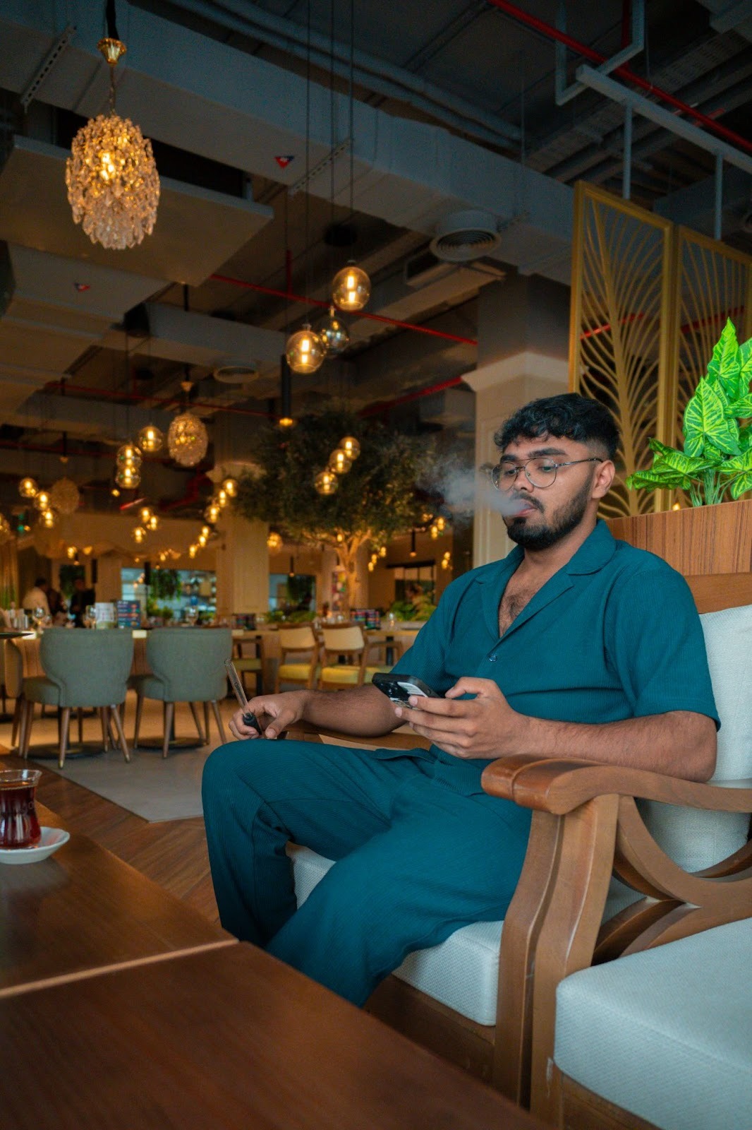 A man sitting comfortably in a stylish, warmly lit lounge, wearing teal scrubs and holding a phone while waiting for his shisha.